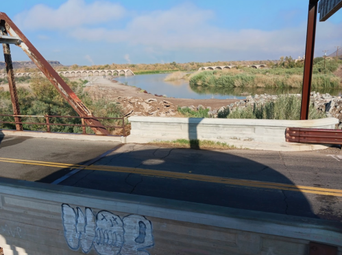 A view of a river with a bridge and overgrown banks under a clear blue sky, featuring graffiti on the foreground.